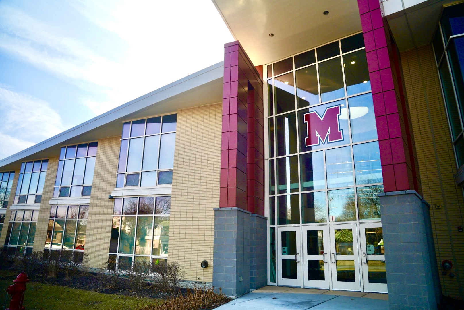 A modern school building featuring tall glass windows, light-colored walls, and a distinctive 'M' logo above the entrance.